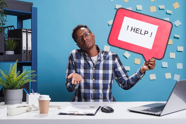 Confused and clueless company office worker holding help cardboard bubble sign while shrugging in workspace. Bewildered uncertain agency employee raising shoulders ambiguous.