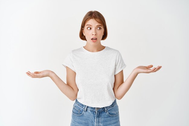 Confused and clueless girl cant understand something looking aside at empty space spread empty hands sideways and shrugging unaware standing on white background