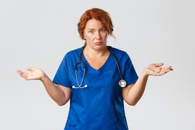 Confused and clueless middle-aged female doctor, physician or nurse in blue scrubs shrugging with concerned look, shaking head, do not know