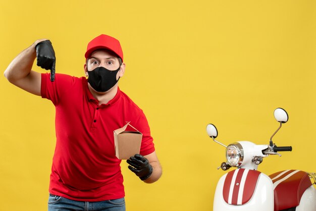 Confused courier man in red uniform wearing black medical mask and glove delivering orders pointing down on white background