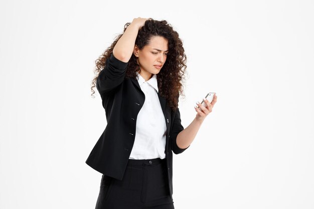 Confused curly business woman holding phone over white wall