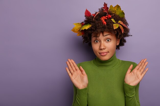 Confused curly female raises palms, gestures in studio, has clueless face expression, says I am not guilty, wears colorful fallen autumn leaves and rowan berries in hairstyle