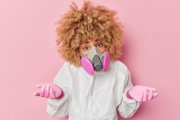 Confused curly haired woman protects herself from chemical poisoning wears safety white suit respirator and gloves shrugs shoulders with bewilderment isolated over pink background Biohazard