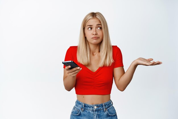 Confused cute blond girl holds mobile phone shrugs shoulers and looks away dont know not understand stands against white background
