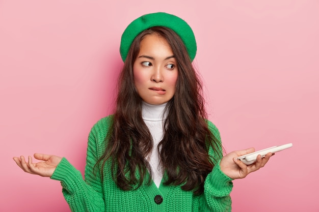 Confused dark haired teenager with asian appearance, holds mobile phone, spreads hands sideways, bites lower lip confusingly, cannot understand how download application, wears green clothes