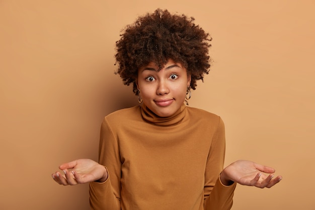 Free photo confused dark skinned adult female has question, feels indecisive and hesitant, shrugs shoulders, poses unaware, being confused, cant help, wears brown turtleneck, isolated on beige wall