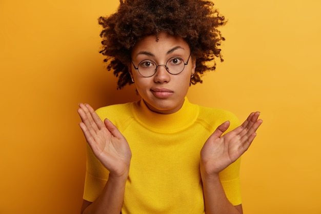 Free photo confused dark skinned female spreads palms and shruggs shoulders, feels indecisive, asks question, doesnt understand what happening, looks through transparent glasses, isolated on yellow wall