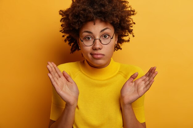 Confused dark skinned female spreads palms and shruggs shoulders, feels indecisive, asks question, doesnt understand what happening, looks through transparent glasses, isolated on yellow wall