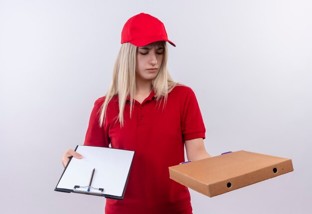 Confused delivery young woman wearing red t-shirt and cap holding clipboard looking pizza box on hand on isolated white wall