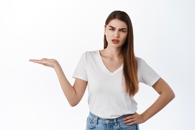 Confused and disappointed girl holding in open hand item showing bad object in her palm display something awful against copy space frowning and complaining white background