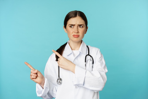 Confused, disappointed medical worker, frowning while pointing fingers left with doubtful, puzzled face expression, standing over torquoise background