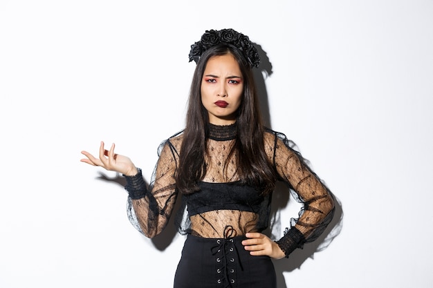 Foto gratuita la donna asiatica confusa e scontenta non può capire qualcosa, alzando la mano e accigliandosi frustrata, indossando l'abito da festa di halloween, in piedi su sfondo bianco.