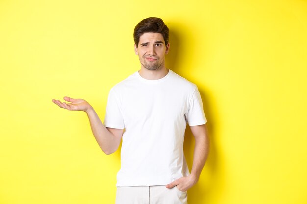 Confused and displeased guy raise hand, grimacing puzzled, standing against yellow background