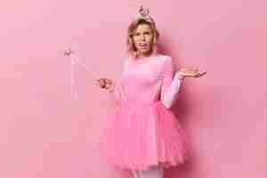 Free photo confused displeased woman shrugs shoulders looks with indignation cannot understand what you meant wears festive dress and magic wand has image of princess on party isolated over pink studio wall