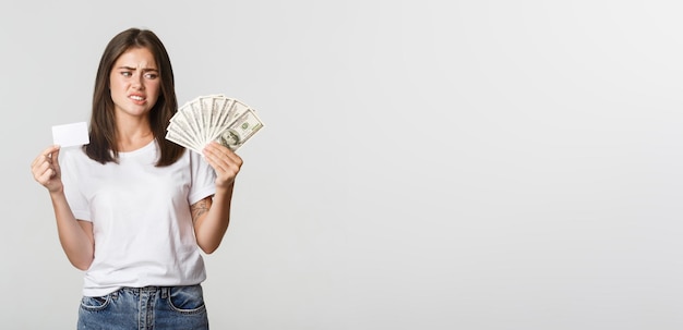 Free photo confused and displeased young woman looking at money and frowning prefer credit card