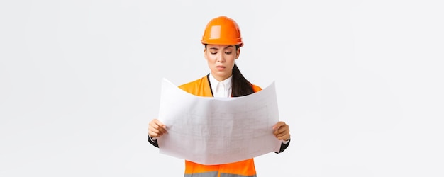 Confused and doubtful asian female chief engineer lead architect at construction area looking displeased and hesitant at strange blueprints reading confusing building project white background