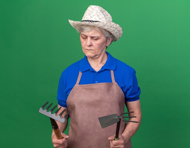 Confused elderly female gardener wearing gardening hat holding rake and hoe rake on green