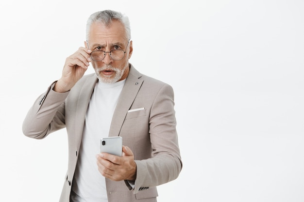 Смущенный пожилой мужчина в костюме держит мобильный телефон и смотрит