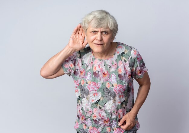 Смущенная пожилая женщина держит руку за ухом, пытаясь услышать, изолированную на белой стене