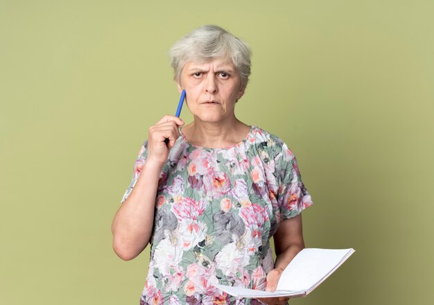 Confused elderly woman holds notebook and puts pen on face isolated on olive green wall