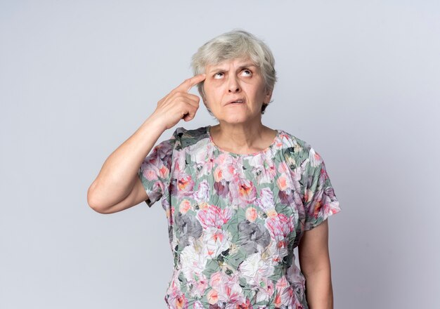 Confused elderly woman puts finger on temple looking up isolated on white wall