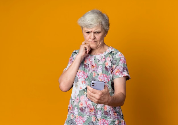 Confused elderly woman puts hand on chin looking at phone isolated on orange wall