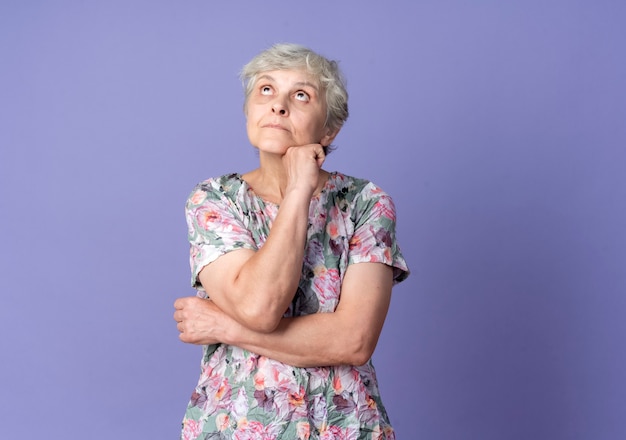 混乱した年配の女性は、紫色の壁に孤立して見上げるあごに手を置きます