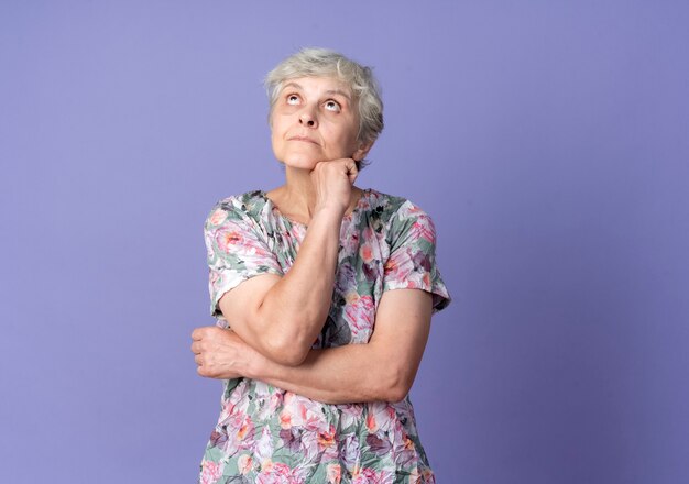 Confused elderly woman puts hand on chin looking up isolated on purple wall