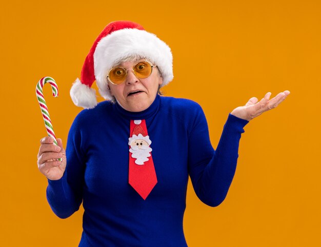 Confused elderly woman in sun glasses with santa hat and santa tie holding candy cane and keeping hand open isolated on orange wall with copy space