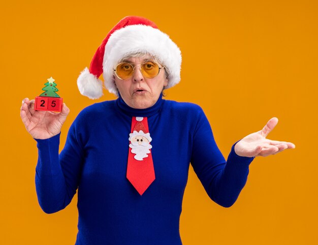 Confused elderly woman in sun glasses with santa hat and santa tie holding christmas tree ornament and keeping hand open isolated on orange background with copy space