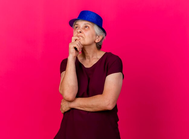 Смущенная пожилая женщина в партийной шляпе держит подбородок, глядя вверх на розовый