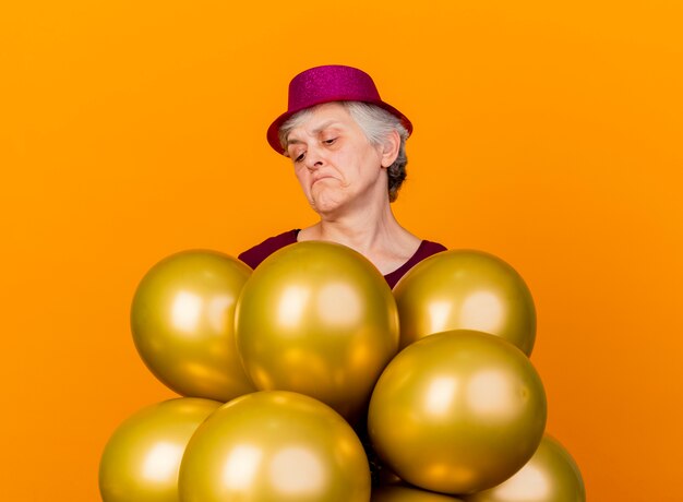パーティーハットをかぶって混乱している年配の女性は、コピースペースでオレンジ色の壁に分離されたヘリウム風船を保持し、見ています