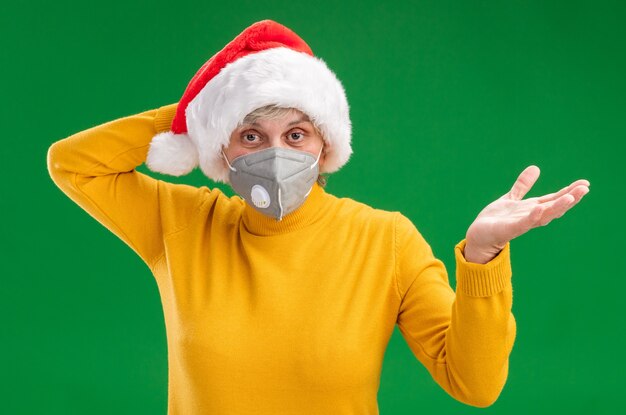 Confused elderly woman with santa hat wearing medical mask puts hand on head and keeps hand open isolated on green wall with copy space