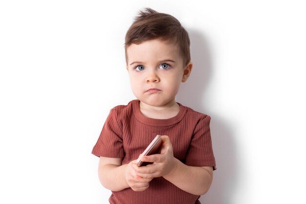 Смущенный эмоциональный взволнованный мальчик-малыш в повседневной одежде, держа смартфон в руках, чувствуя ...