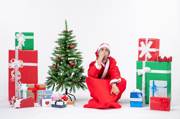 혼란 스 러 워 흥분된 젊은 성인 선물 산타 클로스로 옷을 입고 흰색 배경에 바닥에 앉아 장식 된 크리스마스 트리