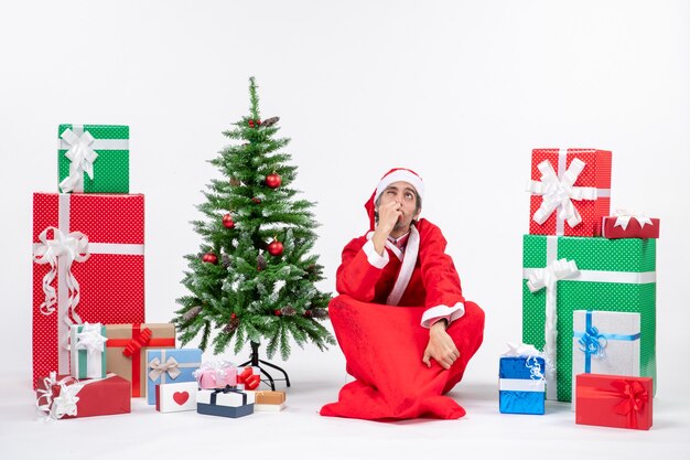 Confused excited young adult dressed as Santa claus with gifts and decorated Christmas tree sitting on the ground on white background
