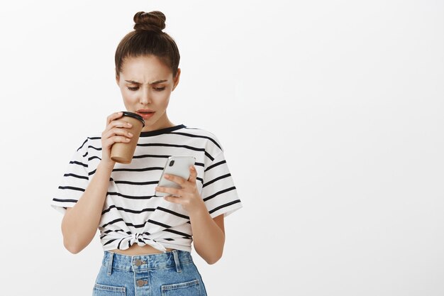 Confused frowning girl looking at mobile phone screen while making sip of coffee from cup