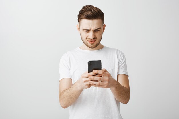 Смущенный и разочарованный парень смотрит на мобильный телефон озадаченно
