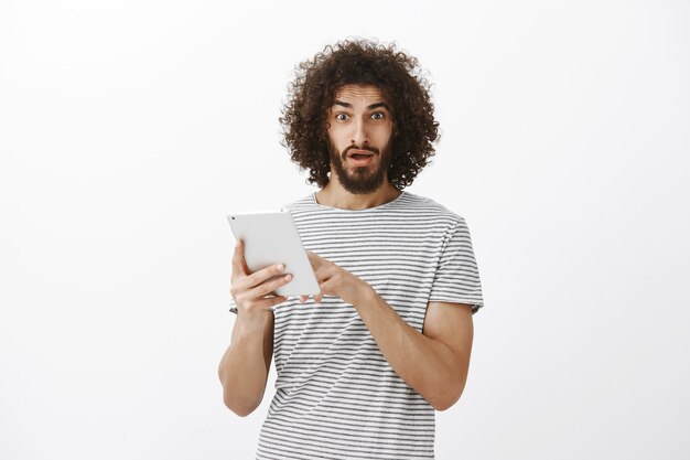 ひげとアフロのヘアカットで欲求不満なハンサムな男を混乱させ、デジタルタブレットの画面を指してあごを落とし、信じられないほどの奇妙なニュースを目にする