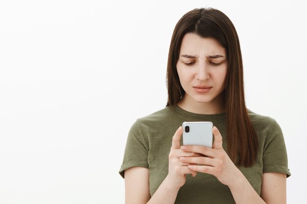 Confused and frustrated young woman cannot understand meaning of message frowning and looking questioned at smartphone screen as reading strage text, posing clueless and focused over gray wall