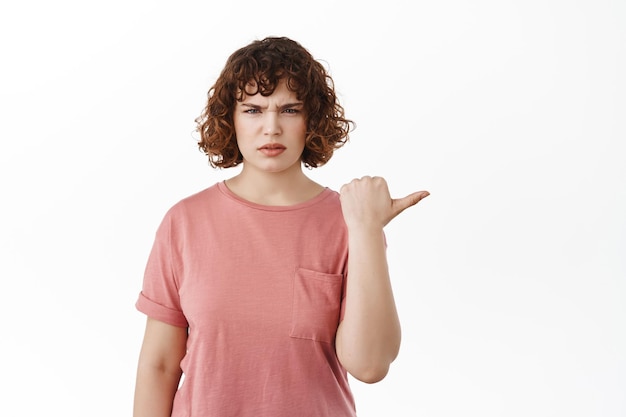 Free photo confused girl pointing right at something strange, frowning and staring displeased or questioned, standing against white background in t-shirt