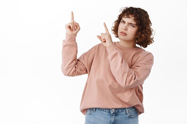 Confused girl points at upper left corner, looks puzzled and clueless, need answers, asking question about advertisement on top, what is this, standing over white background