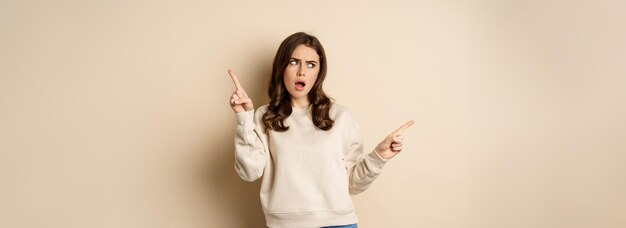 Confused girl shopper pointing sideways making choice decision between two variants beige background