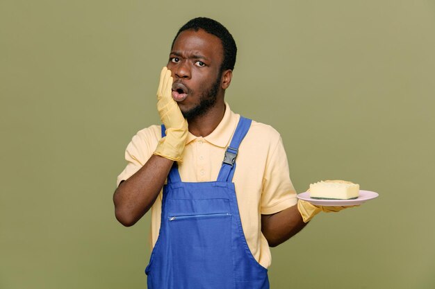 Confused grabbed chin holding sponge on dish young africanamerican cleaner male in uniform with gloves isolated on green background