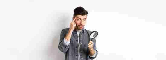 Free photo confused guy found something down with magnifying glass look puzzled at camera standing against whit