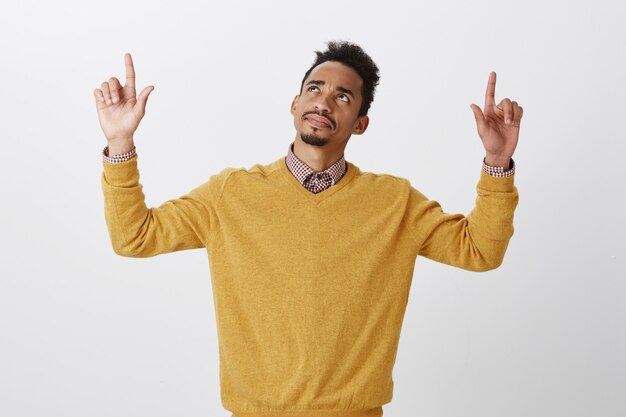 Confused guy, having trouble to recognize something upstairs. Questioned beautiful dark-skinned male model with afro hairstyle, raising hands, pointing and looking up with doubts and suspicion