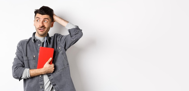 Confused guy scratch head and look aside at logo puzzled holding red diary or planner standing again