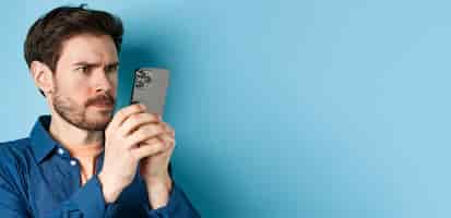 Free photo confused guy staring close at mobile phone screen and frowning standing on blue background