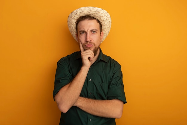 Confused handsome blonde man with beach hat puts hand on chin isolated on orange wall