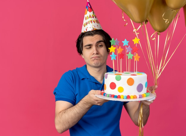 Confused handsome caucasian man wearing birthday cap holds helium balloons and birthday cake 